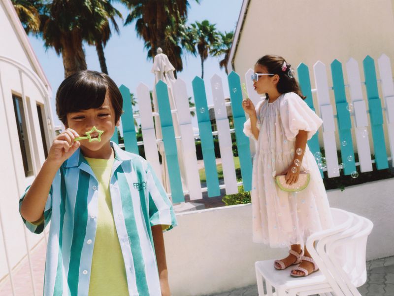 Two children are outside on a sunny day blowing bubbles; a boy in a striped shirt and girl in a dress with a fence backdrop.