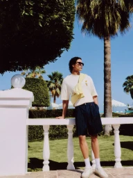 Model leaning on a white balustrade wearing a white t-shirt and dark pinstripe shorts under palm trees