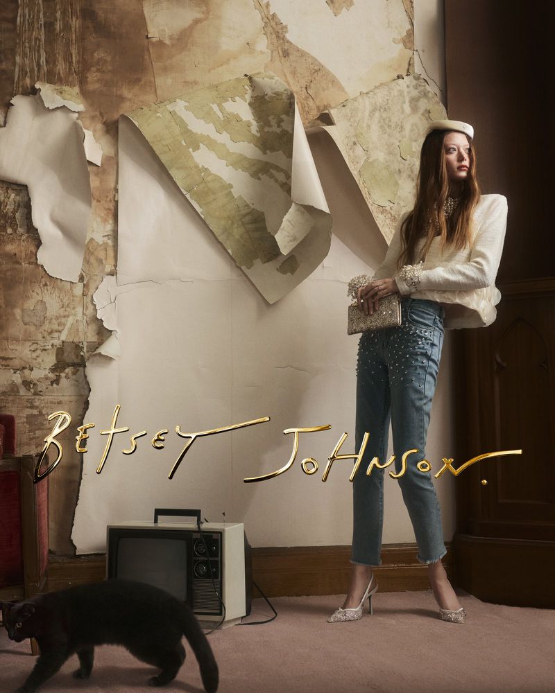 Model Sara Grace Wallerstedt posing in embellished denim, white jacket, and pillbox hat next to a vintage TV and black cat.