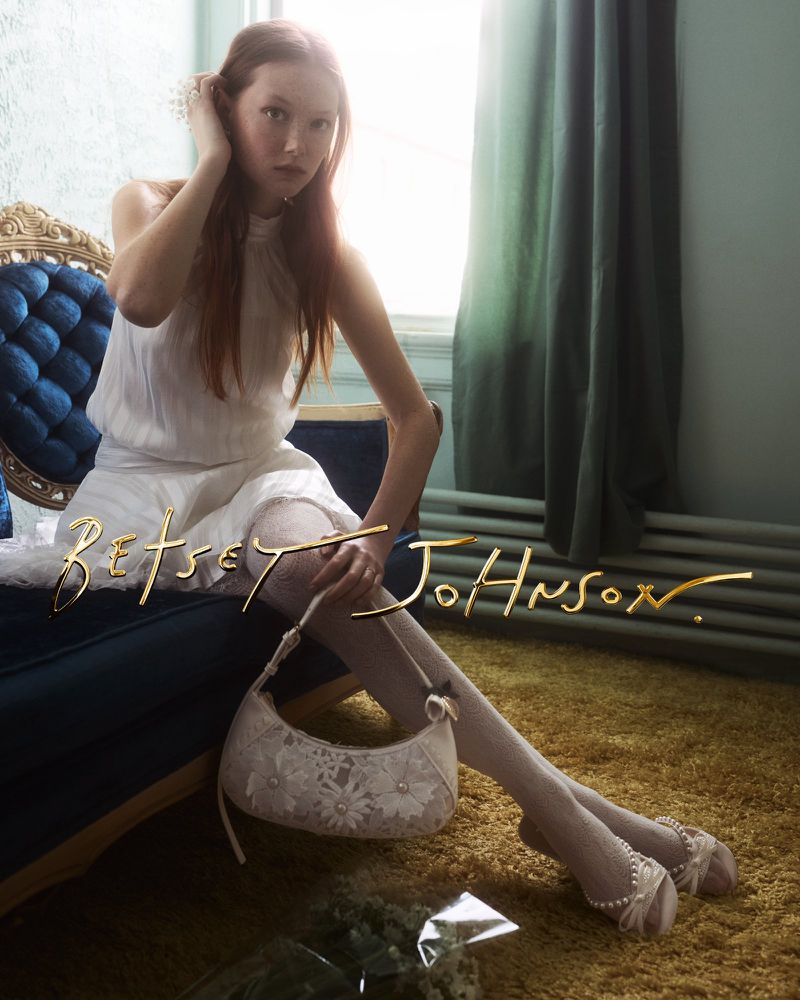 Model sitting on a blue velvet sofa wearing a white dress and holding a patterned handbag in a room with a window
