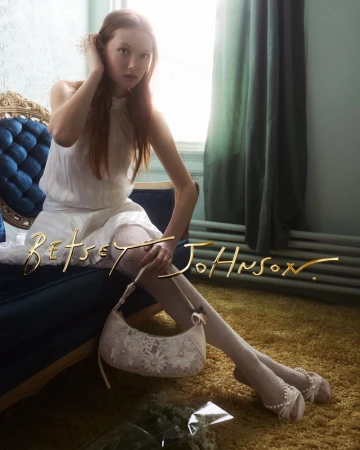 Model sitting on a blue velvet sofa wearing a white dress and holding a patterned handbag in a room with a window