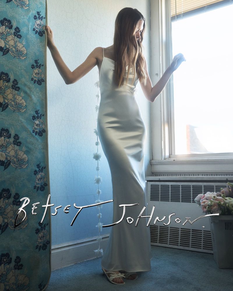 Model Sara Grace Wallerstedt in a white slip dress standing near a window for Betsey Johnson bridal campaign