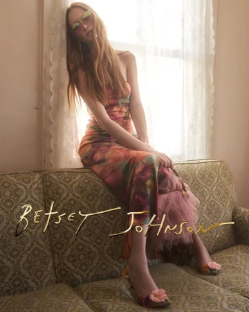 Model Sara Grace Wallerstedt in a floral dress and tinted sunglasses seated on a vintage patterned couch by a lace curtain.