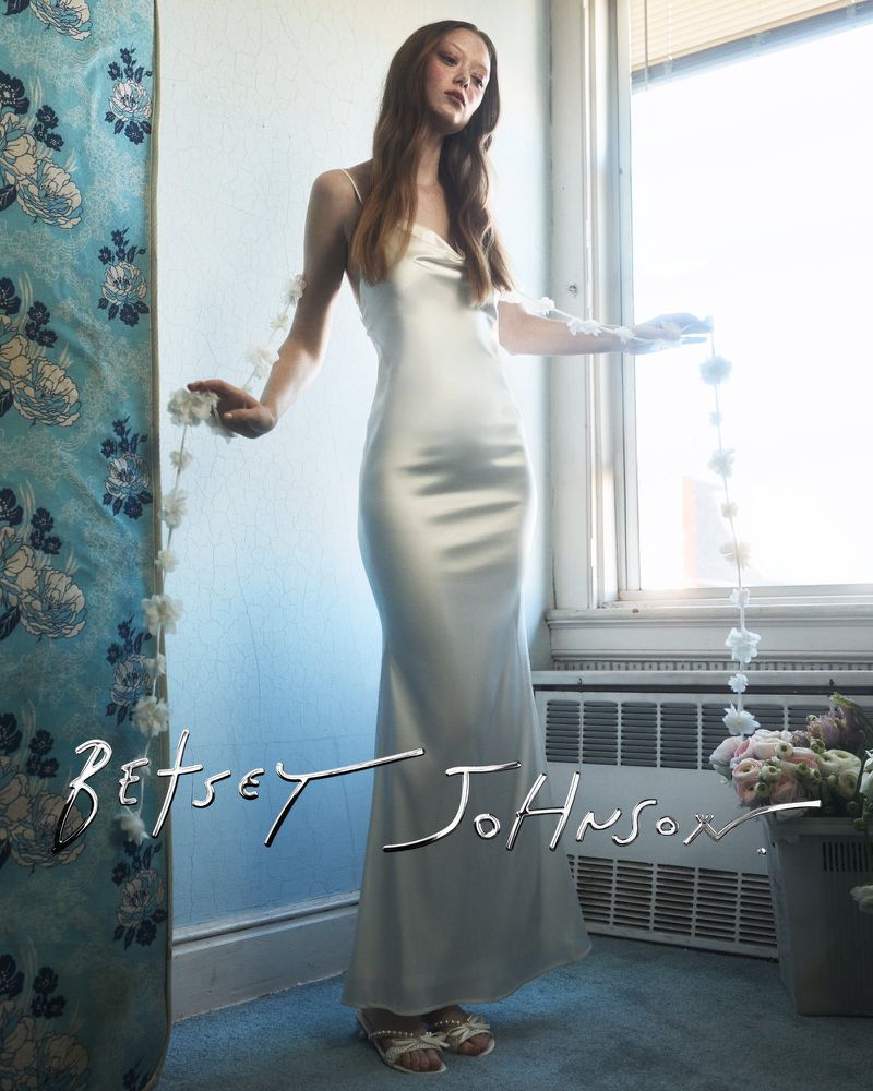 Model in a long satin slip dress holding a floral garland inside a room with a window and blue walls.
