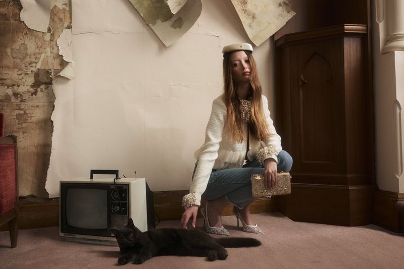 Sara Grace Wallerstedt crouching by a black cat and vintage television in a cream pearl jacket and jeans.