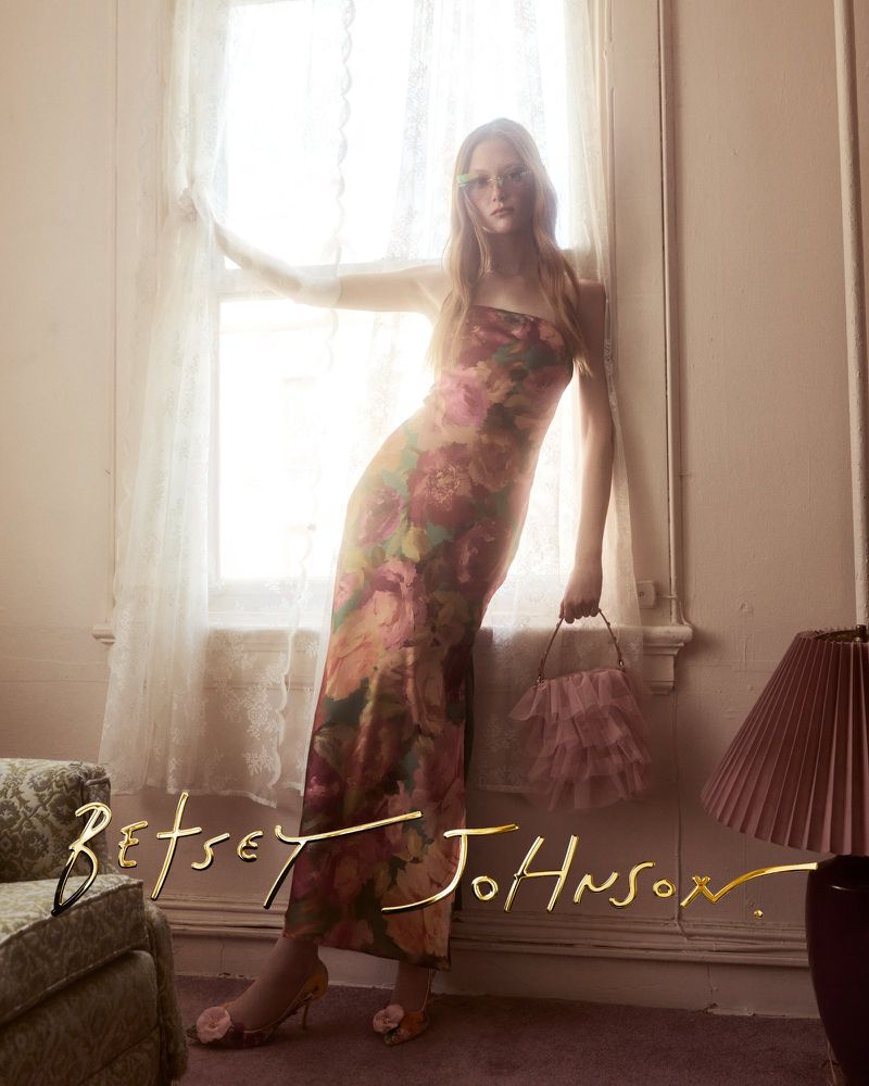 Model Sara Grace Wallerstedt in a strapless floral gown holding a ruffled pink handbag by a lace window.