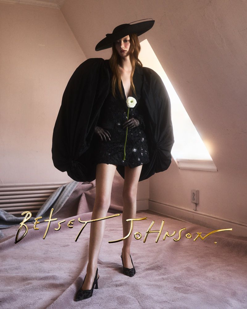 Model Sara Grace Wallerstedt in a black dress, wide-brimmed hat, and oversized sleeves holding a flower in an indoor room