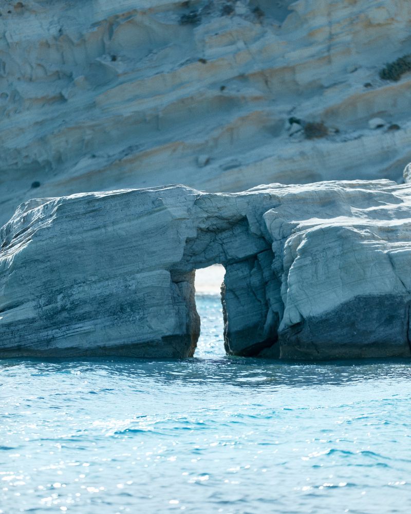 Sea arch formation carved into a light grey and white cliff face along a blue water's edge with clear skies in the background.