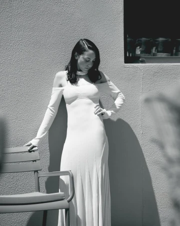 Monochrome portrait of a woman posing in a long white cold-shoulder dress against a textured outdoor wall beside a patio chair.