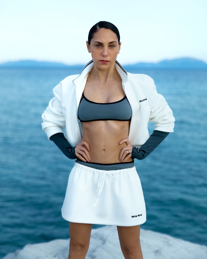 Athletic woman in a white jacket, grey sports bra, and white skirt poses outdoors on a stone surface with a sea and sky background.