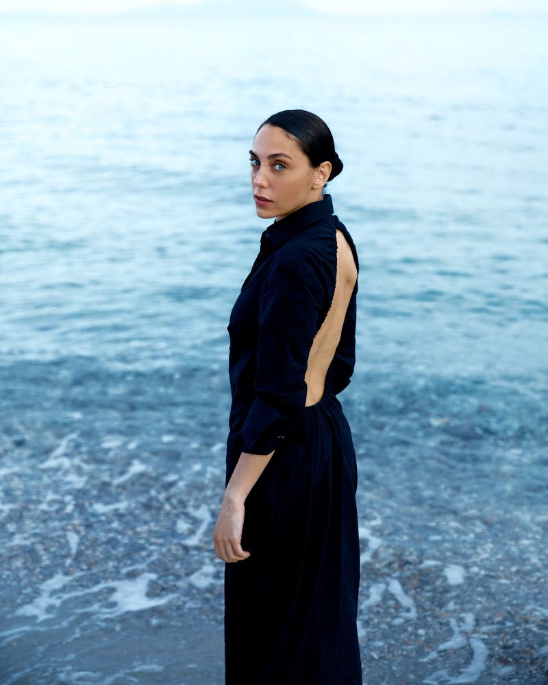 Woman in black dress stands on a beach with ocean waves in the background. She is looking over her shoulder at the camera.