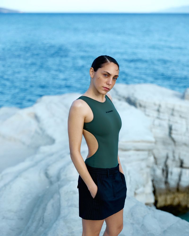 Woman wearing a dark green top and black skirt posing on light rocky coastal shore against blue ocean water.