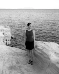 Woman standing on rocky coastal cliffs wearing a dark sleeveless top and skirt with the ocean in the background