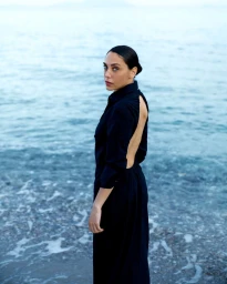 Woman in an open-back black long-sleeved dress looking over her shoulder while standing at the ocean shoreline.