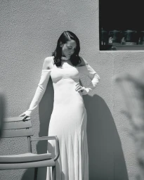 Monochrome portrait of a woman posing in a long white cold-shoulder dress against a textured outdoor wall beside a patio chair.