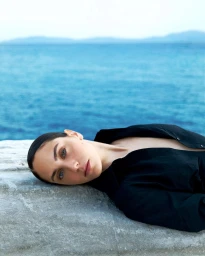 Woman in an open black shirt reclining on a stone ledge with a calm blue ocean in the background.