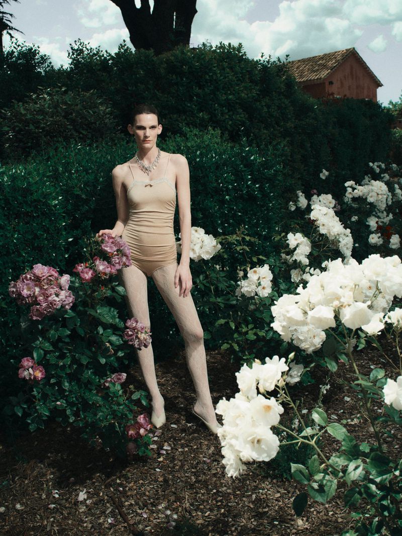 Model Lena Hardt wearing a nude bodysuit and matching tights stands amidst blooming white and mauve rose bushes in an outdoor garden.