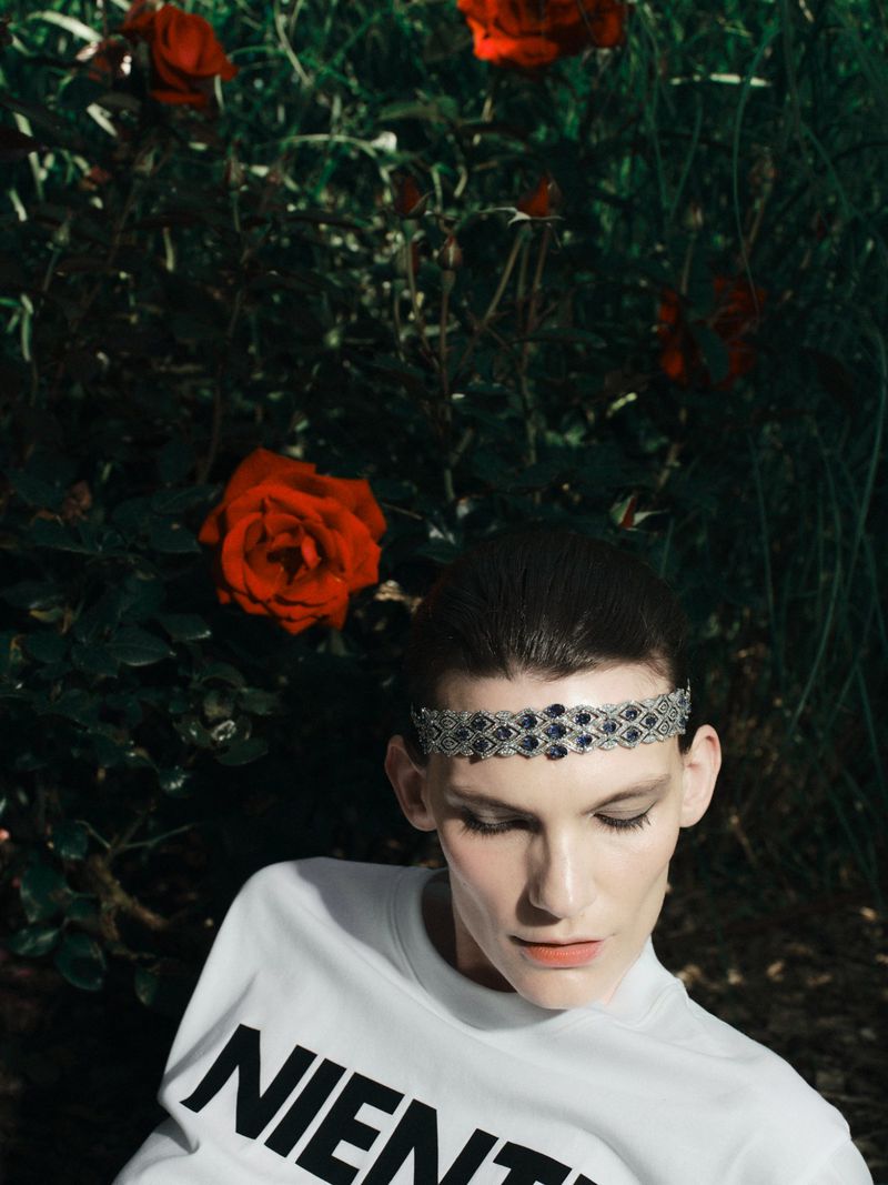 Model wearing a beaded headband and white graphic sweatshirt posing in front of dark green foliage with red roses