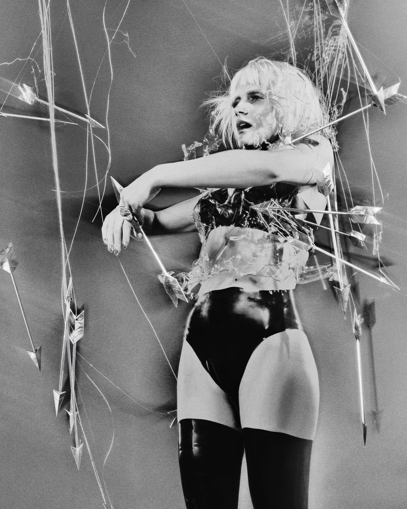 Mala Emde posing in black latex briefs and a clear sculpted bodice surrounded by suspended arrows against a studio backdrop.