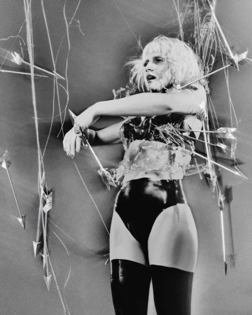 Mala Emde posing in black latex briefs and a clear sculpted bodice surrounded by suspended arrows against a studio backdrop.