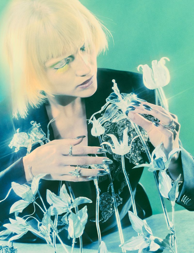 Blonde woman wearing a black jacket examining delicate glass flower sculptures for Vogue Germany editorial.
