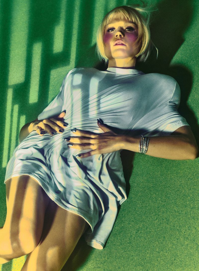 Model with blonde bob wearing a light grey dress lying on a green surface patterned with yellow light stripes