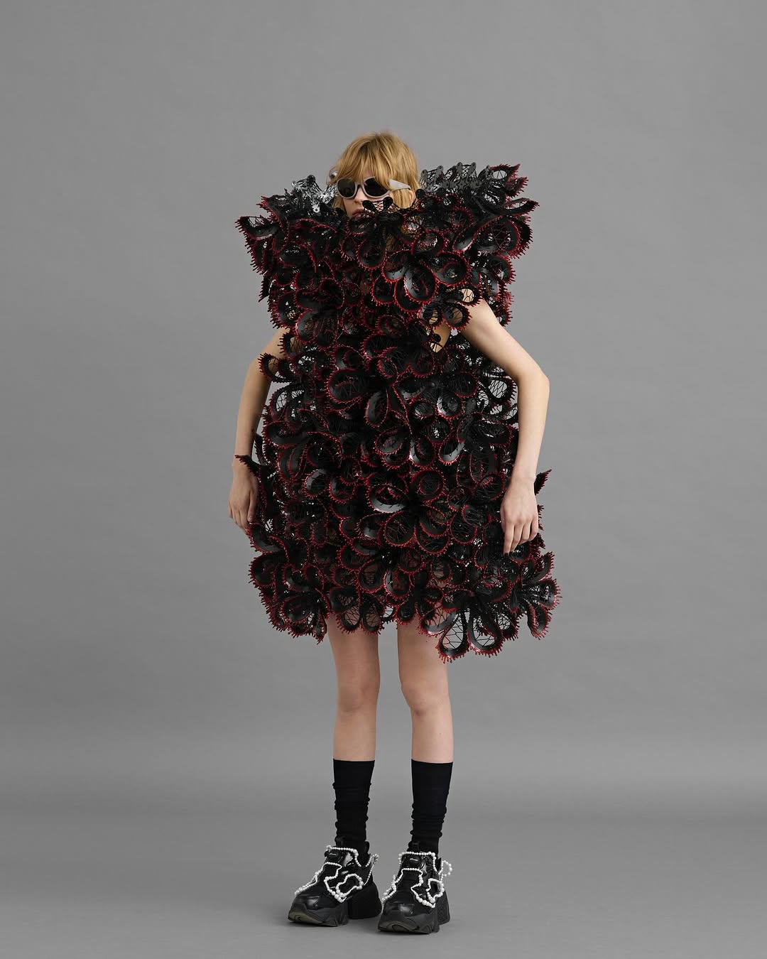 A person with light blonde hair wearing sunglasses and a black and red frilled dress stands against a gray backdrop.