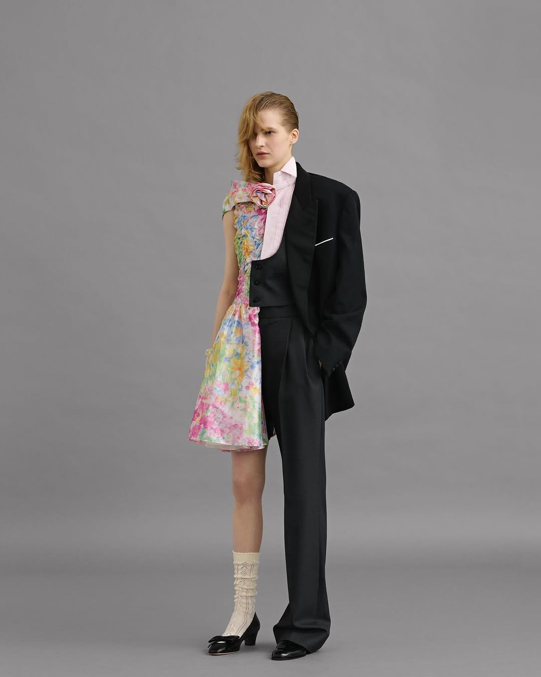 Woman in half floral dress and half black tuxedo stands in a photo studio with a serious expression, showcasing fashion and style.