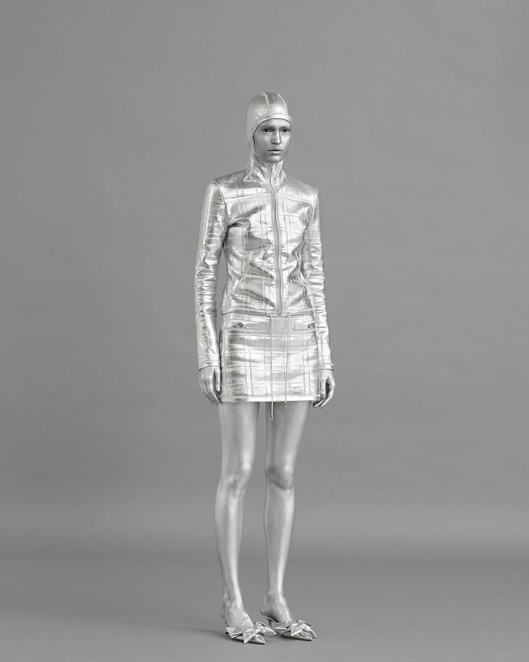 Silver-painted woman in matching mini skirt and jacket with hood poses in a fashion-style studio shot on a gray backdrop.