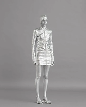 Silver-painted woman in matching mini skirt and jacket with hood poses in a fashion-style studio shot on a gray backdrop.