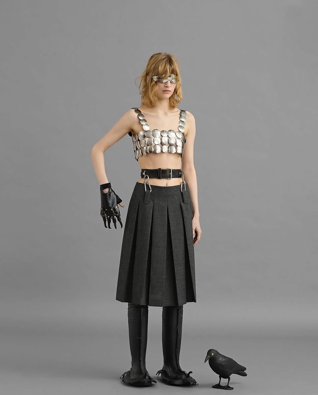 Full shot of a young adult woman posing in a metal-disc top, pleated skirt, and black boots, standing next to a black raven on grey.