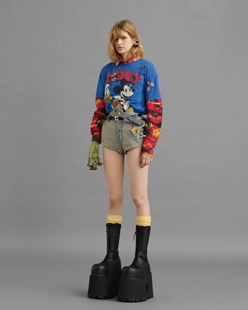 Fashion model poses in studio wearing a graphic sweatshirt, denim shorts, platform boots. Eye-level shot on neutral grey background.