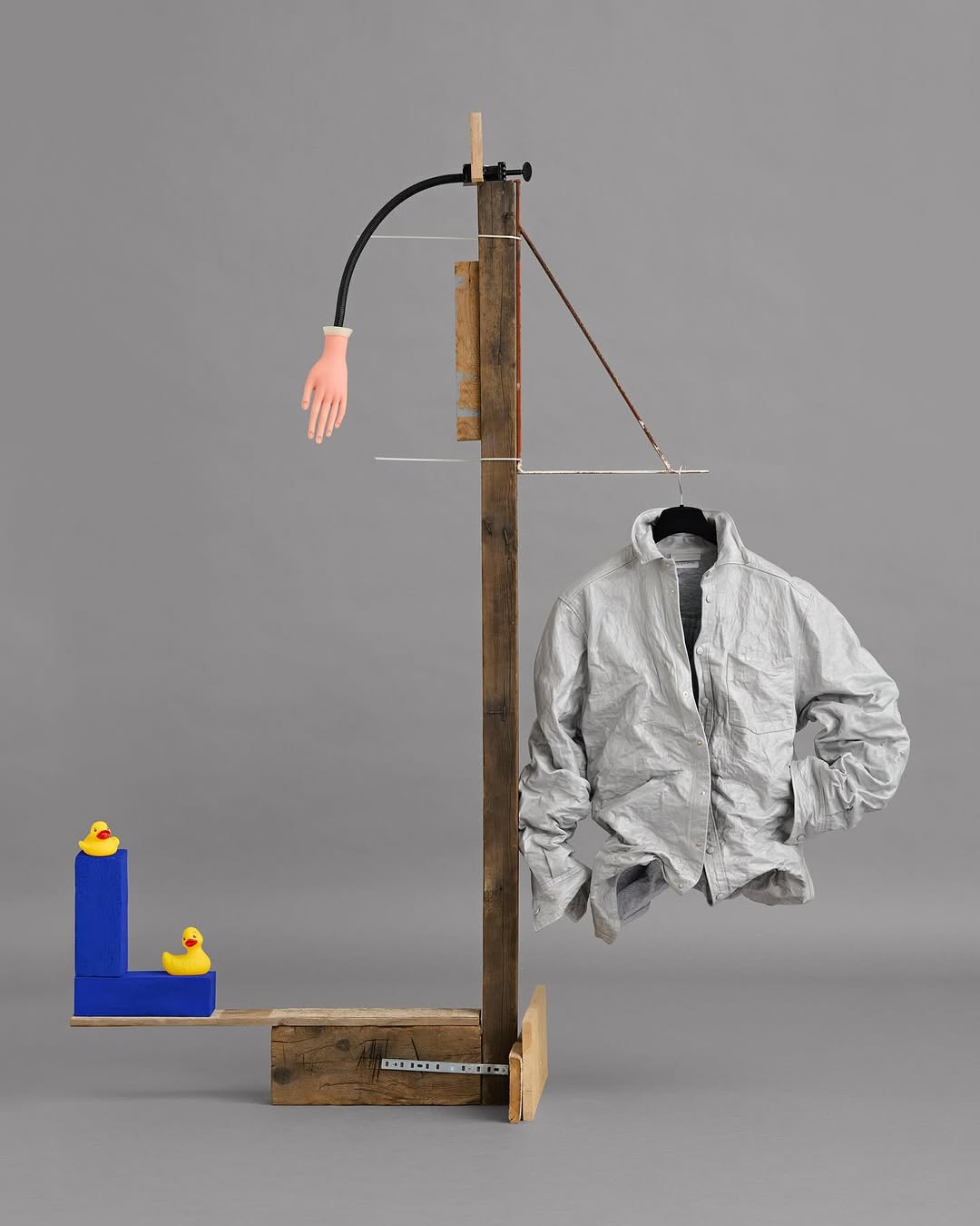 Surreal composition with an off-white jacket on a hanger, a wooden stand, a rubber hand, and toy ducks against a grey background.