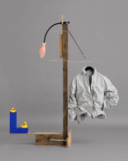 Surreal composition with an off-white jacket on a hanger, a wooden stand, a rubber hand, and toy ducks against a grey background.