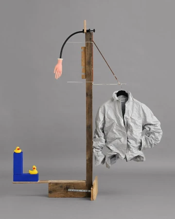 Surreal composition with an off-white jacket on a hanger, a wooden stand, a rubber hand, and toy ducks against a grey background.