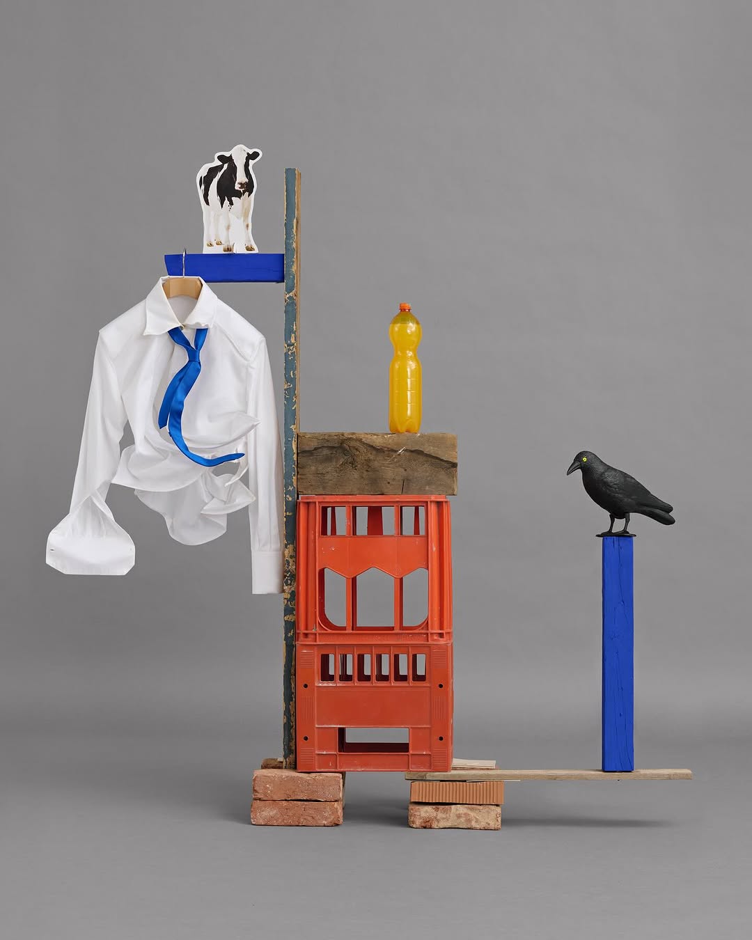 Artistic still life: white dress shirt on hanger, orange crates, yellow bottle, and black crow figurine on grey background.