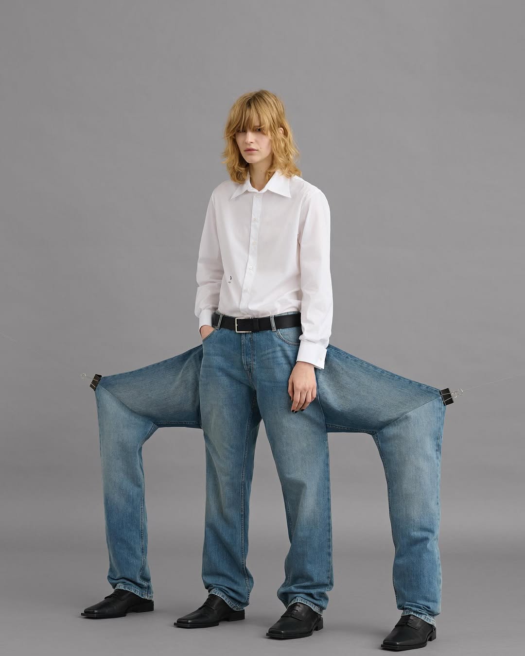 Model wearing a white shirt and avant-garde multi-legged blue jeans against a grey seamless background.