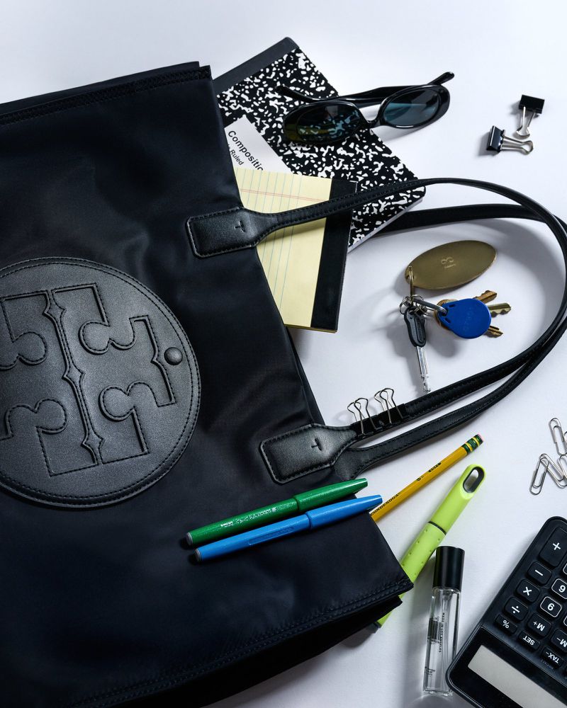 Tory Burch black tote bag with sunglasses, notebook, pens, keys, and calculator against a white background in a studio setting.