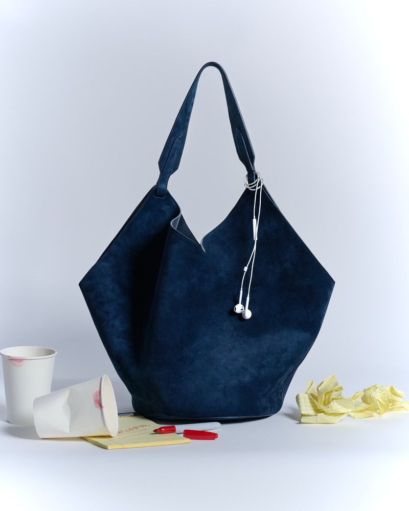 Dark blue suede tote bag with white earbud headphones, surrounded by crumpled paper, pens, sticky notes, and paper cups with lipstick marks.