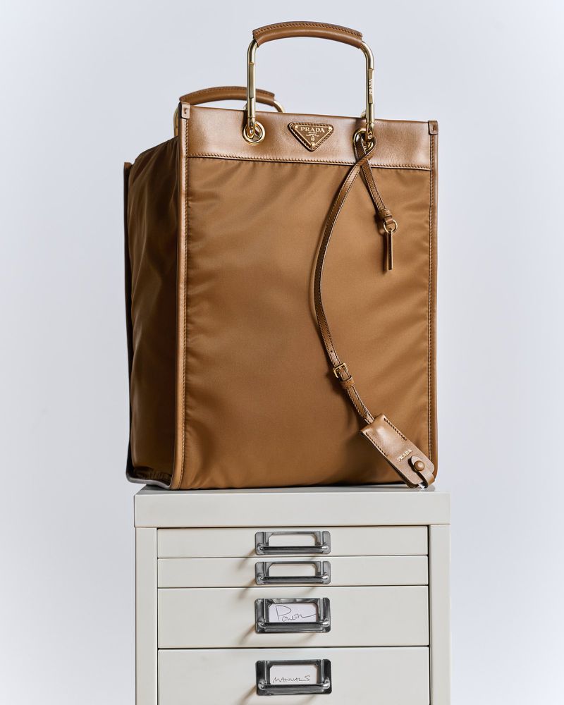 A brown Prada handbag with gold hardware and leather trim sits atop a white filing cabinet against a neutral studio backdrop.