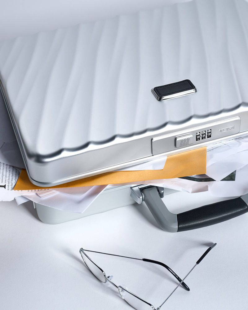 Open metal briefcase with combination lock and spilled documents, accompanied by a pair of glasses, all set against a plain background.