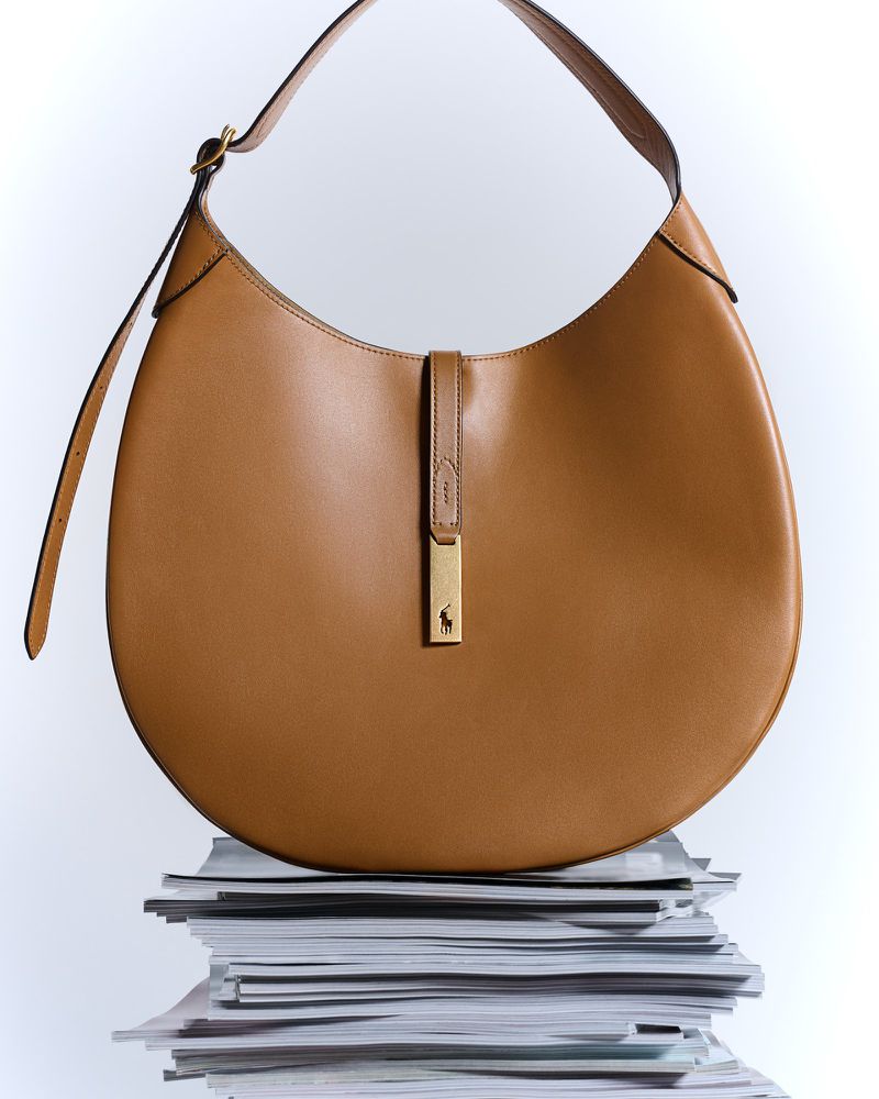Tan leather hobo bag with gold hardware and a logo detail, placed on a stack of magazines against a white backdrop in a studio shot.