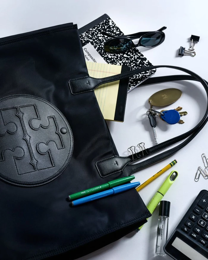 Top-down flat lay of a black designer tote bag surrounded by stationery, sunglasses, keys, and a calculator on a white surface.