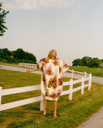 Model Georgina Grenville wearing an oversized floral cocoon dress with arms outstretched beside a white fence in a rural meadow.