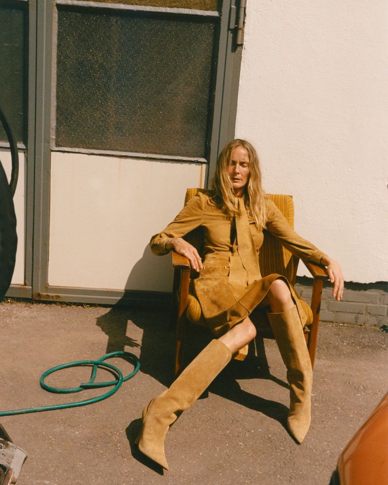 Model seated in an armchair wearing a camel suede dress and tall boots outside against a building exterior.
