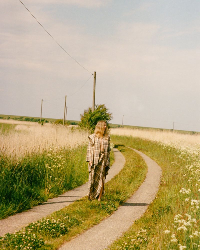 Model Georgina Grenville walking along a rural dirt road surrounded by grassy meadow fields in a plaid coat outfit.