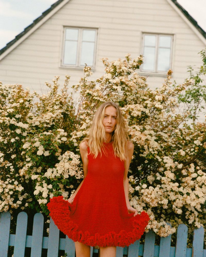 Model Georgina Grenville poses in a red knit ruffle-hem mini dress in front of flowering bushes and a blue fence.