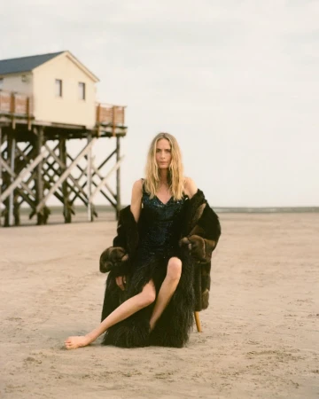 Georgina Grenville seated barefoot on a sandy beach wearing a dark sequined dress and fur coat in front of a stilt house.