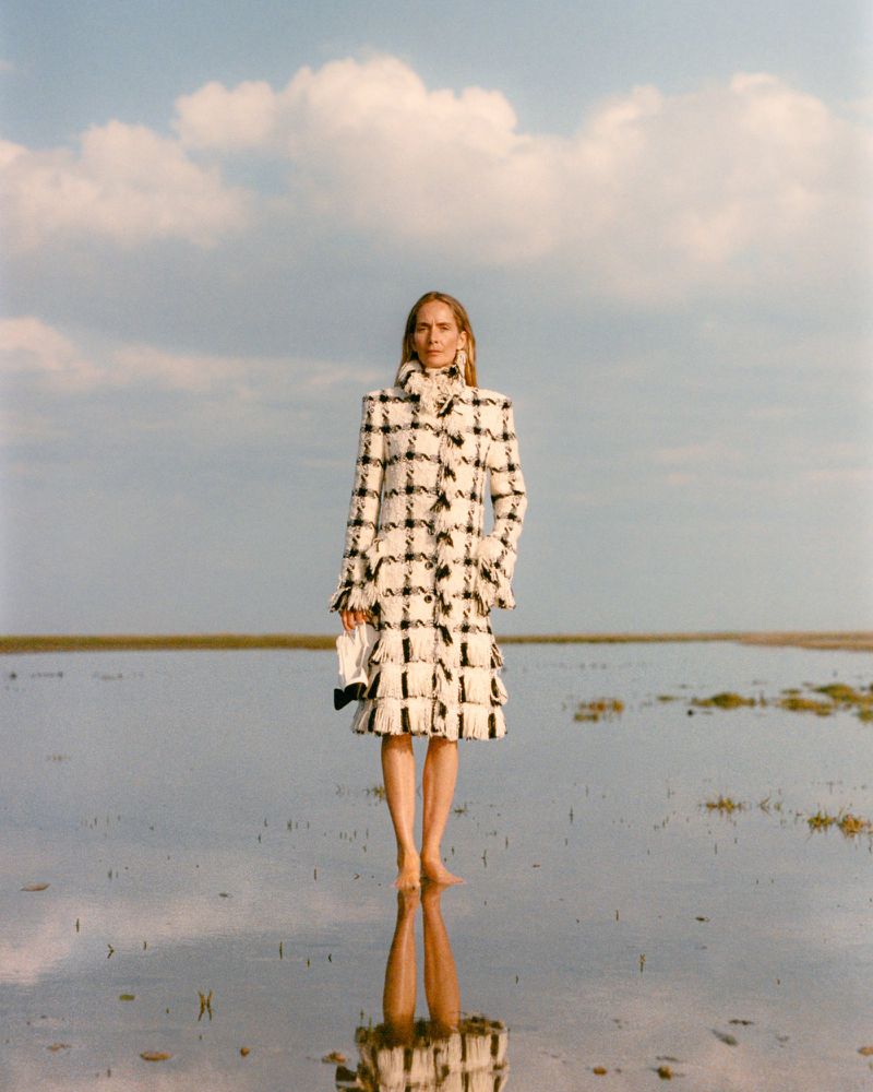 Model Georgina Grenville in a black and white fringe plaid coat standing barefoot in shallow reflective water under a cloudy sky.