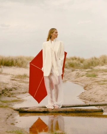 Georgina Grenville standing barefoot on a log in an ivory blazer holding a red geometric umbrella in coastal sand dunes.